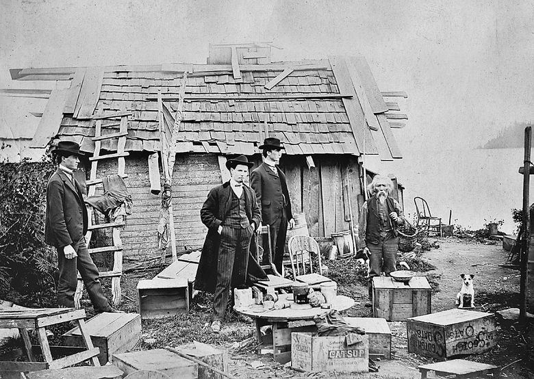 Salmon Bay Charlie, at right, with a clam basket over his arm and a dog at his side, is photographed with three suited men as he is evicted from his home. The site is near the present-day West Sheridan Street-end Park in Magnolia. (Photo by Clarence Langstaff, circa 1900. Courtesy of the Magnolia Historical Society Archive)