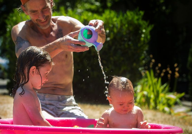 Jason Ludwick plays with his granddaughters in a tiny pool in front of his un-air-conditioned home in Des Moines last week. At left is Cherish Rain Jimenez, 2, and at right is Trinity Nicole Joy Jimenez, 9 months. (Ellen M. Banner / The Seattle Times)
