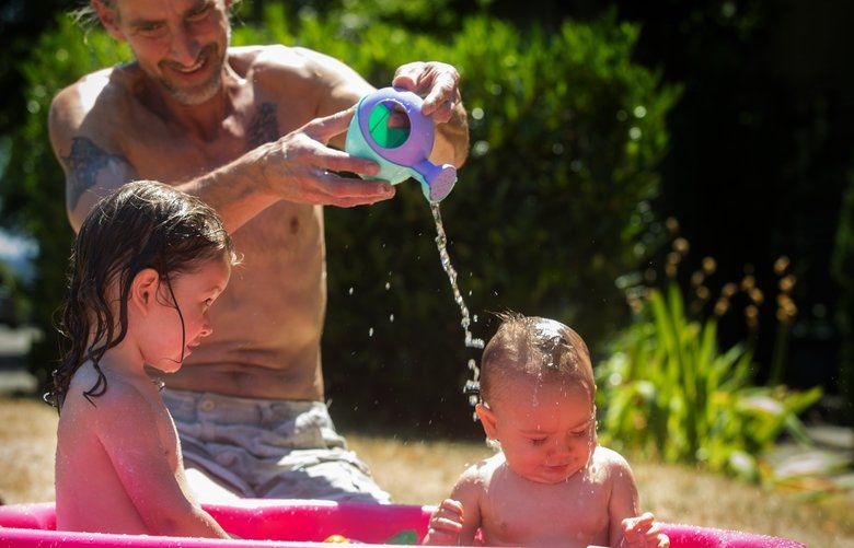 Jason Ludwick (c) plays with his granddaughters in a tiny pool in front of his Des Moines home Monday, July 16, 2018.  At left is Cherish Rain Jimenez, 26 mos., and at right is Trinity Nicole Joy Jimenez, 9 months.  He and his wife watch the children while their mother attends classes at Highline College.  Temperatures in the high 80’s are expected Tuesday and in the mid to high 70’s the rest of the week.