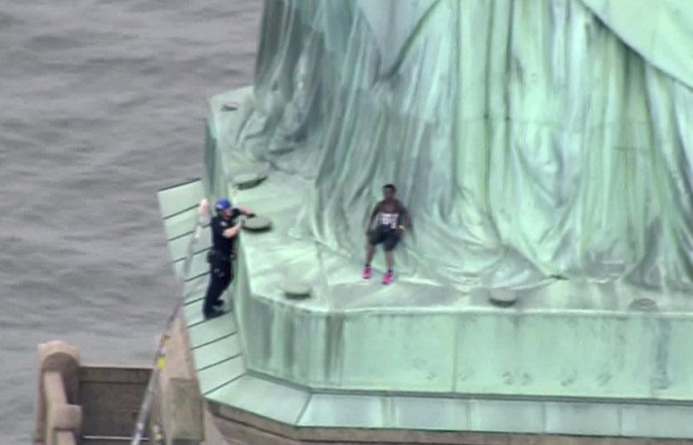 In this image made from video by PIX11, a person, center, leans against the robes of the Statue of Liberty on Liberty Island, as one of the police officers climbed up on a ladder to stand on a ledge nearby talking the climber into descending in New York, Wednesday, July 4, 2018. (PIX11 via AP) NYHK302 NYHK302