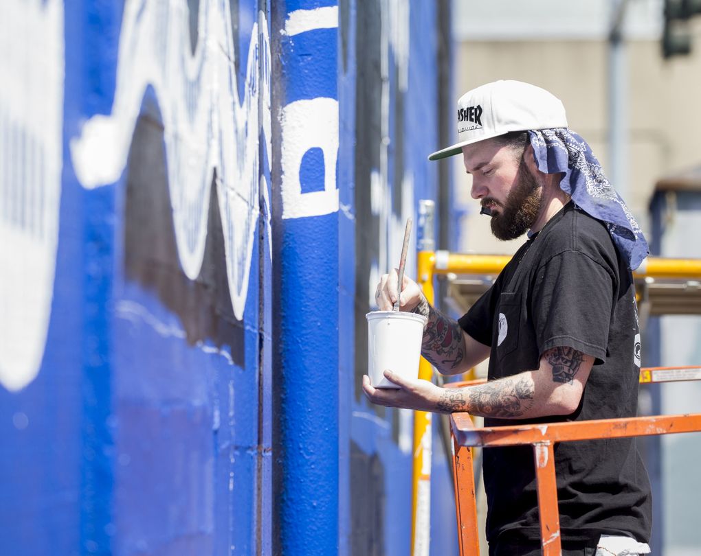 Seattle artist Jesse Brown paints his mural, a study in blue and black of shapes, snakes, eyes, keyholes and sunbursts, over another painting he’d made there 10 years ago. (Bettina Hansen/The Seattle Times)