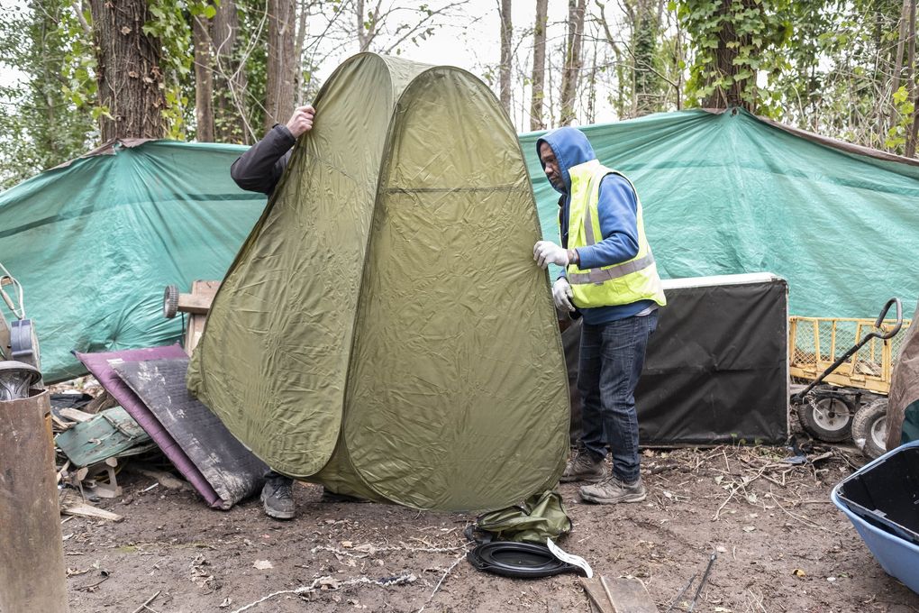 Eric Smith sets up a privacy screen for use as a toilet in the homeless encampment he lives in near Rainier Avenue. It was given to him by Mark Lloyd, left, who has provided some basic elements of sanitation few homeless sites have. Smith, who is homeless, does what he can to keep his encampment clean. Both he and Lloyd are worried about sanitation in the camps in light of the coronavirus outbreak. (Dean Rutz / The Seattle Times)