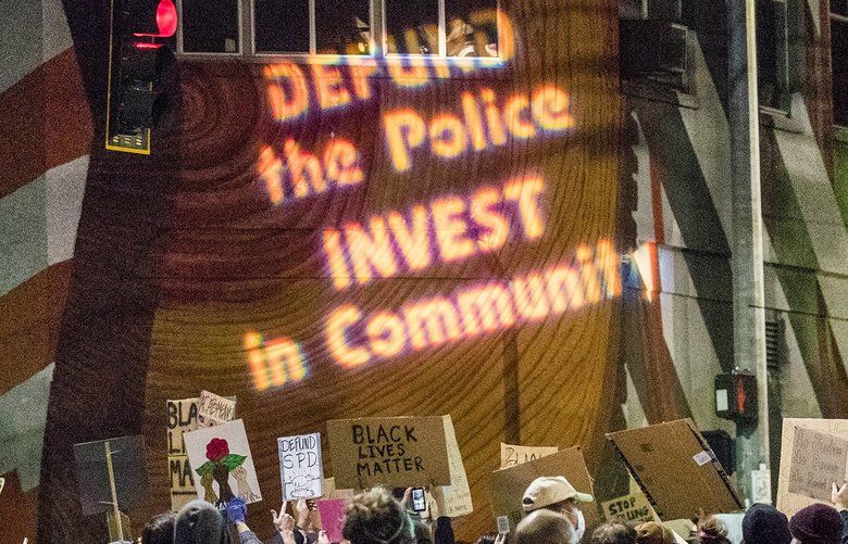 “DEFUND the Police INVEST in Community” is projected on the wall near protesters and police at Pine Street and 11th Avenue on Thursday, June 4, 2020. People wheeled in a stage light on a cart to project messages of solidarity on the wall.  214165 214165