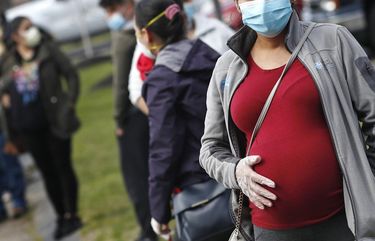 FILE – In this Thursday, May 7, 2020 file photo, a pregnant woman wearing a face mask and gloves holds her belly as she waits in line for groceries with hundreds during a food pantry sponsored by Healthy Waltham for those in need due to the COVID-19 virus outbreak, at St. Mary’s Church in Waltham, Mass. On Thursday, June 25, 2020, the Centers for Disease Control and Prevention updated its list of which Americans are at higher risk for severe cases of COVID-19, adding pregnant women and removing age alone as a factor. (AP Photo/Charles Krupa) NY664 NY664