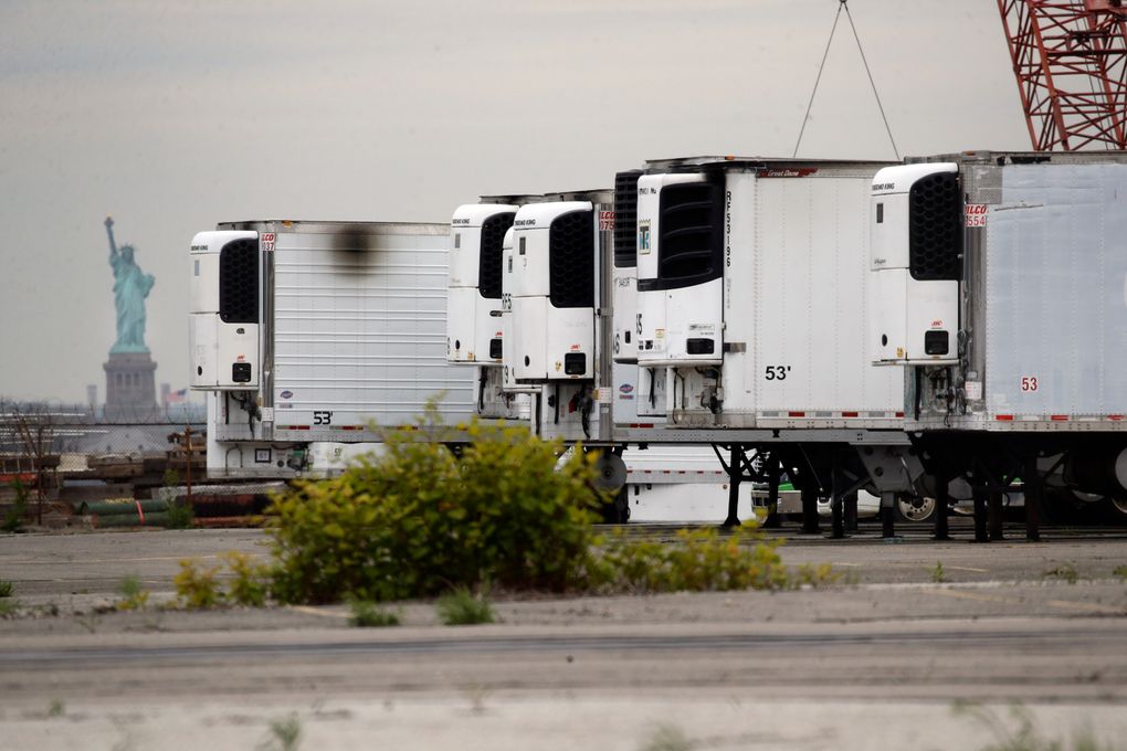 FILE — In this May 6, 2020 file photo, the Statue of Liberty is visible behind refrigerator trucks intended for storing corpses that are staged in a lot at the 39th Street pier, in the Brooklyn borough of New York. New York City is still using refrigerated trucks to store bodies of coronavirus victims, more than a year after they were first set up as temporary morgues as deaths surged at at the height of the pandemic. (AP Photo/Ted Shaffrey, File)