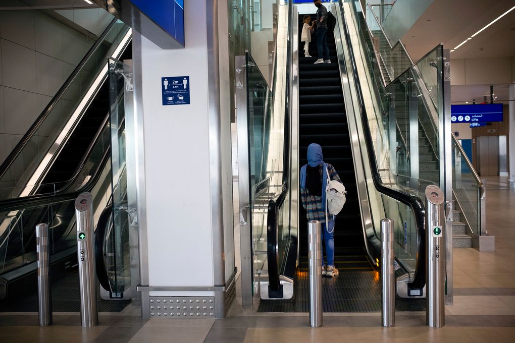 Rahima Asadi hesitates at the bottom of an unfamiliar escalator as her husband, Afghan Air Force Maj. Naiem Asadi, and their daughter, Zainab, look back at her at the airport in Dubai, United Arab Emirates, on June 1, 2021. (Kiana Hayeri / The New York Times)