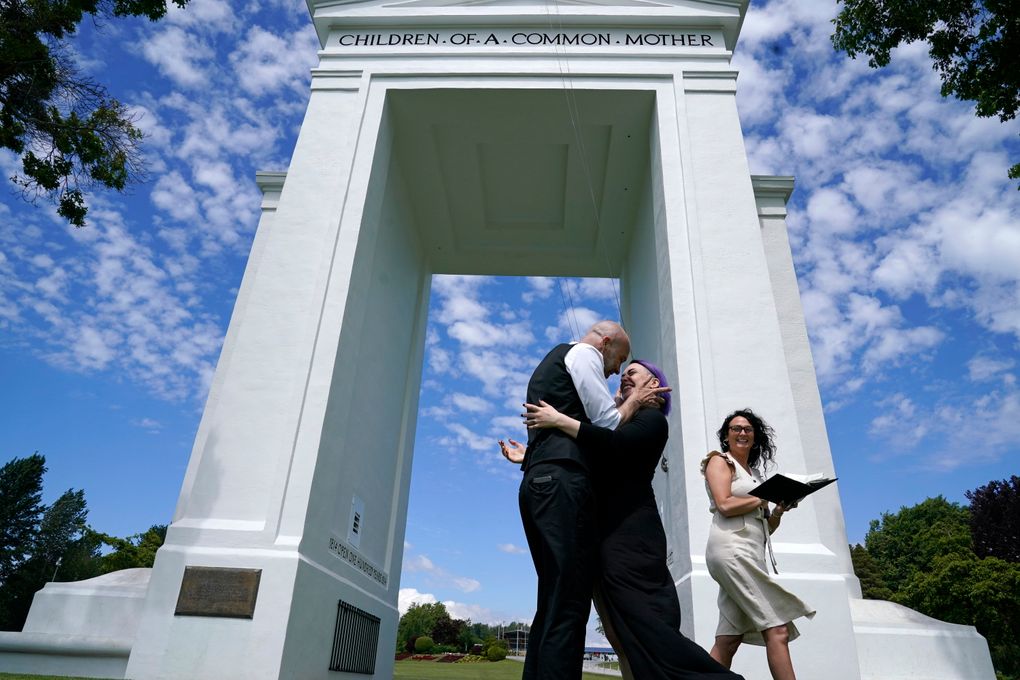 Joel Gardiner, left, embraces his bride, Mandi Gardiner, as officiant Brooke Bakogeorge steps away after marrying the couple at the arch separating the U.S. from Canada at Peace Arch Historical State Park Tuesday, June 8, 2021, in Blaine, Wash. Gardiner, of Canada, walked across the border at the park into the U.S. earlier in the day to wed his American sweetheart. The border has been closed to nonessential travel since March 2020, but Canadians have been allowed to walk over a ditch into the U.S. park and weddings have become routine there. (AP Photo/Elaine Thompson)