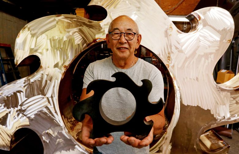 Gerard Tsutakawa, accomplished Pacific NW sculptor, holds the presentation model for “SeaWave,” with the actual bronze piece behind him, still in progress, Wednesday, Aug. 18, 2021 in Seattle. The sculpture, seen in Tsutakawa’s home studio, will be placed outside Climate Pledge Arena, on the International Fountain side. 217958