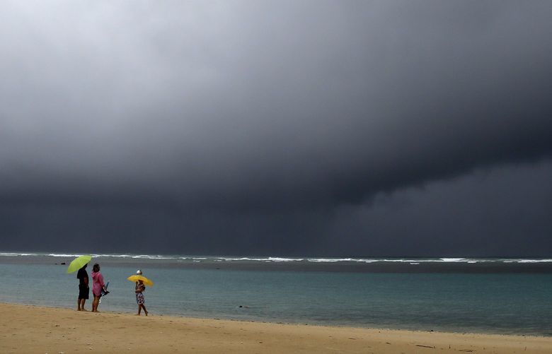 People hold umbrellas as it begins to rain on an otherwise empty beach in Honolulu on Monday, Dec. 6, 2021. A strong storm packing high winds and extremely heavy rain flooded roads and downed power lines and tree branches across Hawaii, with officials warning Monday of potentially worse conditions ahead. (AP Photo/Caleb Jones) HICJ101 HICJ101