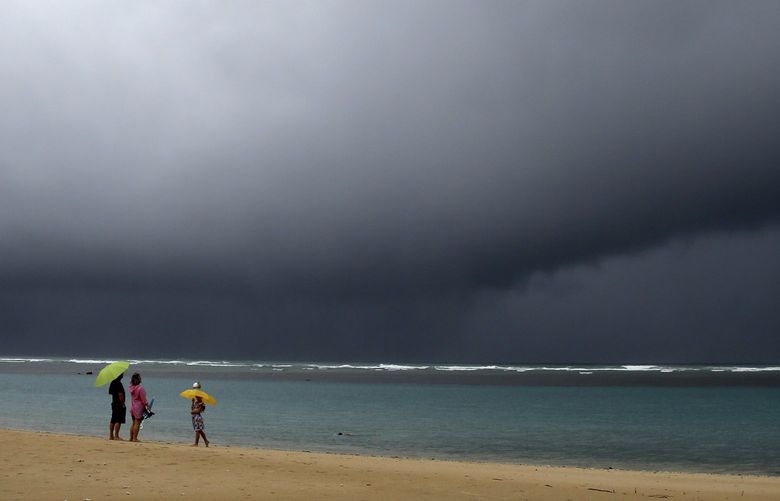 People hold umbrellas as it begins to rain on an otherwise empty beach in Honolulu on Monday, Dec. 6, 2021. A strong storm packing high winds and extremely heavy rain flooded roads and downed power lines and tree branches across Hawaii, with officials warning Monday of potentially worse conditions ahead. (AP Photo/Caleb Jones) HICJ101 HICJ101