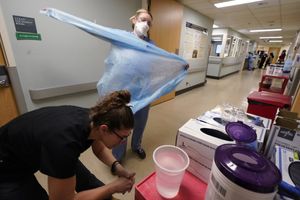 Registered nurse Jessalynn Dest, left, removes protective equipment and washes her hands after leaving a COVID-19 patient’s room as speech therapist Sam Gibbs puts on safety clothing while preparing to see a patient in the acute care unit of Harborview Medical Center earlier this month. (Elaine Thompson / The Associated Press)