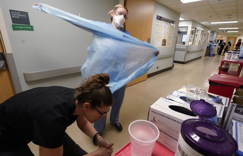 Registered nurse Jessalynn Dest, left, removes protective equipment and washes her hands after leaving a COVID-19 patient’s room as speech therapist Sam Gibbs puts on safety clothing while preparing to see a patient in the acute care unit of Harborview Medical Center, Friday, Jan. 14, 2022, in Seattle. Washington Gov. Jay Inslee is deploying 100 members of the state National Guard to hospitals across the state amid staff shortages due to an omicron-fueled spike in COVID-19 hospitalizations. Inslee announced Thursday that teams will be deployed to assist four overcrowded emergency departments at hospitals in Everett, Yakima, Wenatchee and Spokane, and that testing teams will be based at hospitals in Olympia, Richland, Seattle and Tacoma. (AP Photo/Elaine Thompson) WAET101 WAET101