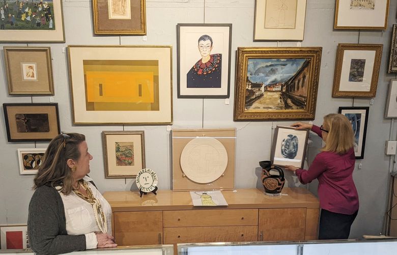 In this image from video, Elizabeth Haynie Wainstein, right, hangs up a piece of modern art belonging to the late Justice Ruth Bader Ginsburg inside Potomack Company Auctions in Alexandria, Va., Monday, April 11, 2022. (AP Photos/Nathan Ellgren) WX103 WX103