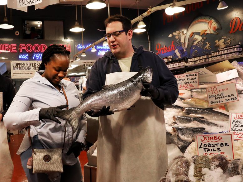 Joydie Ellis came to Pike Place Market to take a souvenir photograph at Pure Food Fish Market’s stand, where co-owner Isaac Behar suggested adding a king salmon to the photo. While businesses in downtown Seattle have seen an uptick in tourists, sales to office workers have not rebounded to pre-pandemic levels. (Alan Berner / The Seattle Times)