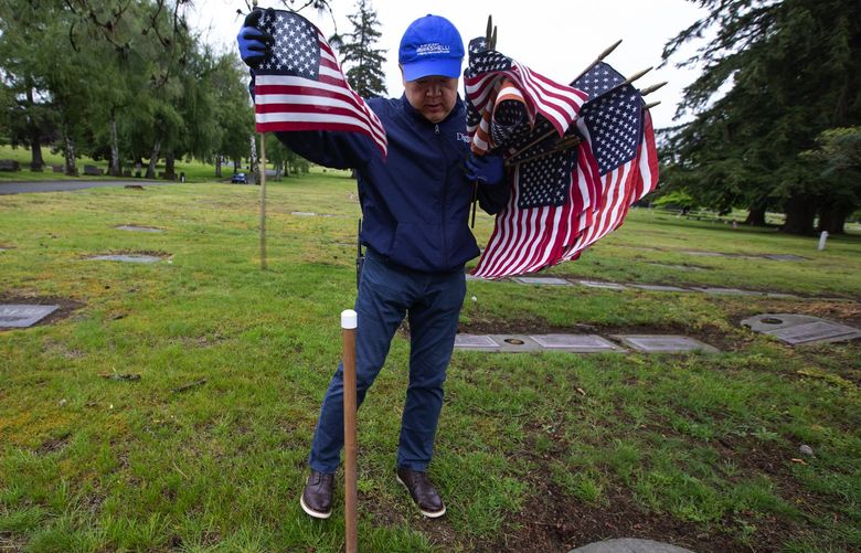 Evergreen Washelli Cemetery employee Keith Lee looks for signs of military service on grave markers to plant U.S. flags in the “traditional” section, which is east of the “memorial park” section that has the uniform upright tombstones, Sunday May 29, 2022 in preparation for Memorial Day in Seattle. At 10 a.m. on Monday, flags will be planted at each upright grave marker in the memorial section, which runs along Aurora Ave., with a service following at 11.