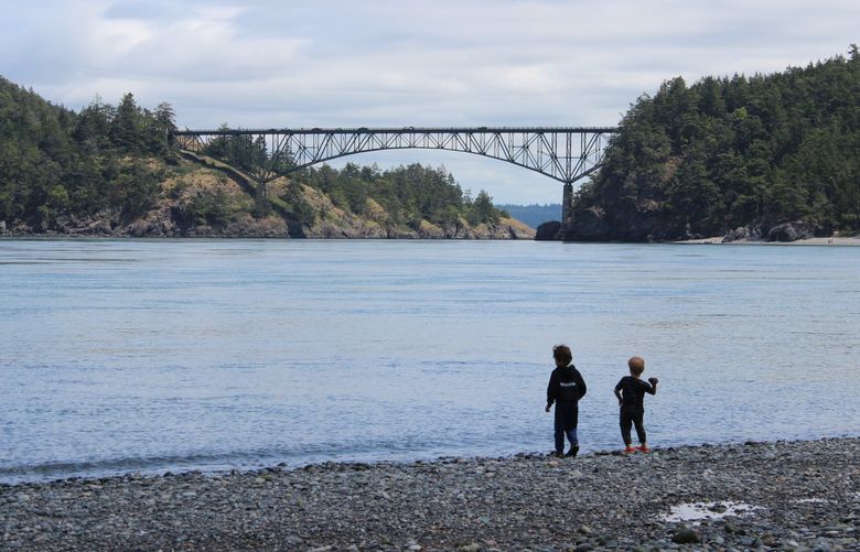 Two children throw works in the water at North Beach with a view of Deception Pass Bridge in the background.