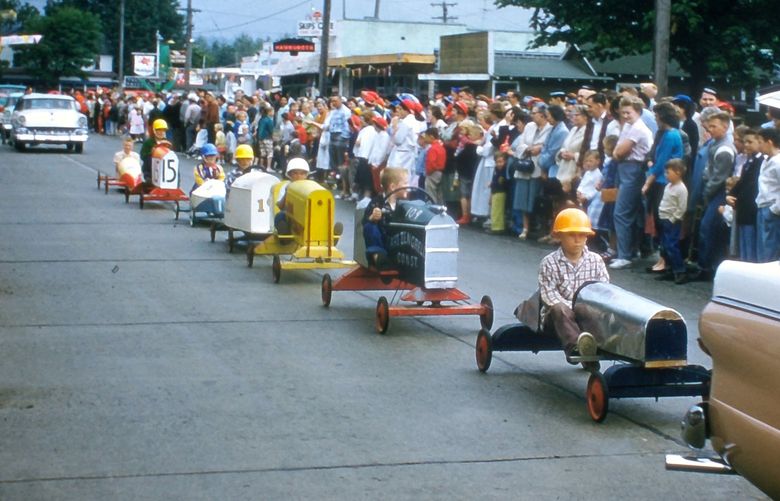 THEN2: A procession of seven Soap Box Derby cars is towed on the same route in the 1956 parade. The Fall City Hotel and Café’s neon sign glows, with the initial letter in the bottom word alternating to drive home the message “GOOD” and “FOOD.” Credit: Larry Divers / Courtesy Fall City Historical Society