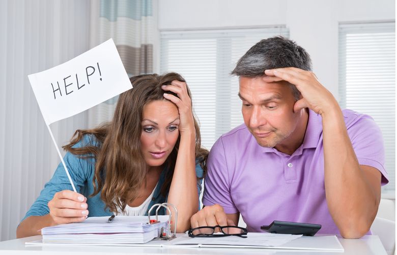A couple sit at a desk looking at their finances—the woman holds up a sign that says "help!"