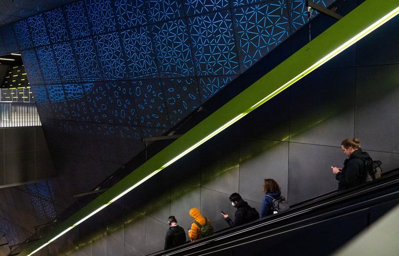 Commuters ride the escalator down to the University of Washington light rail station platform, passing through “Subterraneum,” an art installation created by Leo Saul Berk, on Thursday, Nov. 3, 2022. The light patterns represent the layers of soil that surround the station walls.