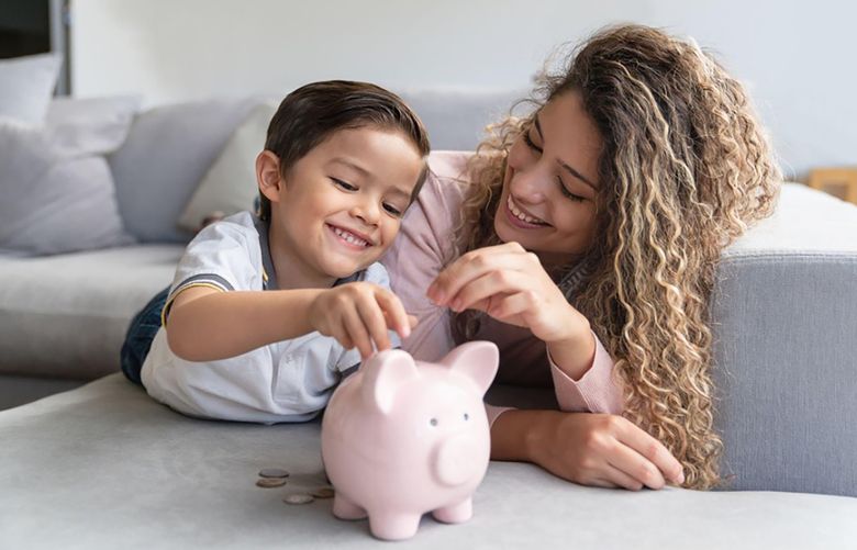 A mother and her son lay on the floor in a living room and place a coin in a piggybank