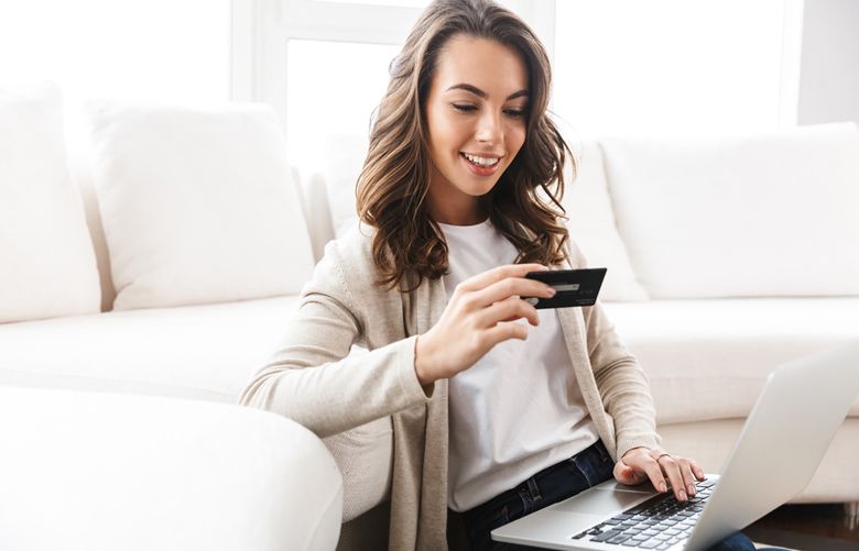 A woman sits in her living room against a cream couch and looks at her credit card in her hand. A laptop is open, resting in her lap.
