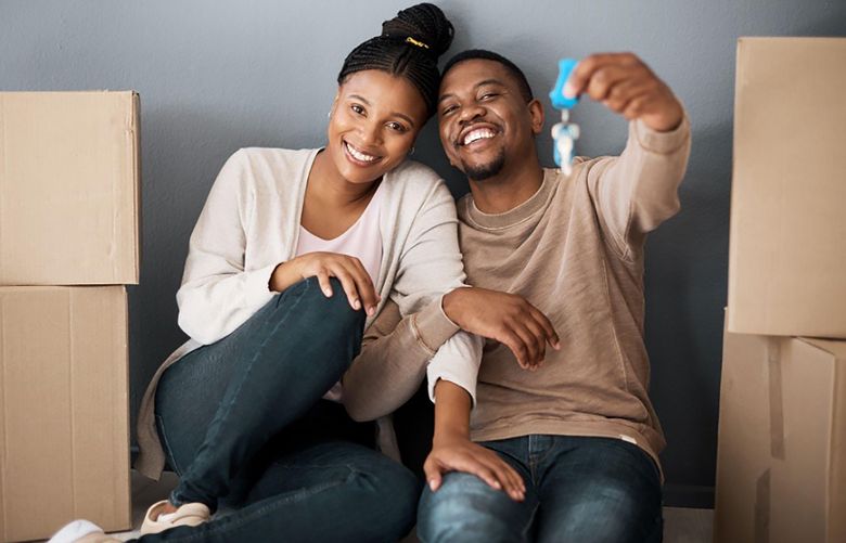 A couple sits against a gray wall amongst moving boxes and holds up a key to their new home happily.