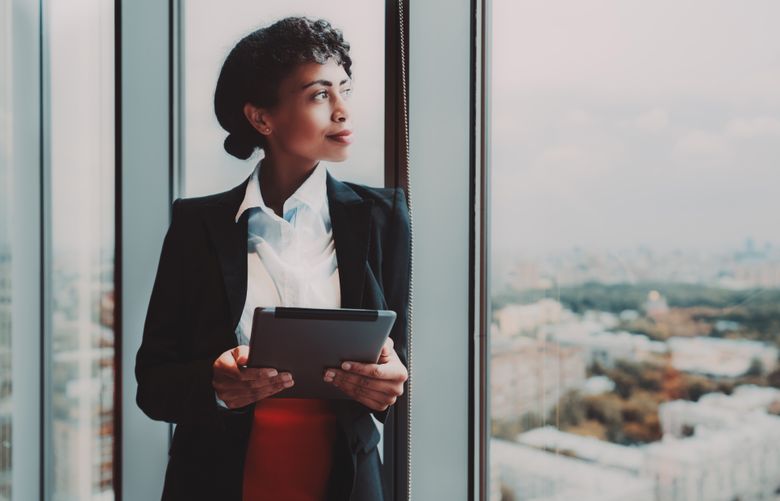 A dazzling young African-American woman entrepreneur with a digital tablet in her hands is standing near a huge panoramic window indoors of a business high-rise and pensively looking at the cityscape