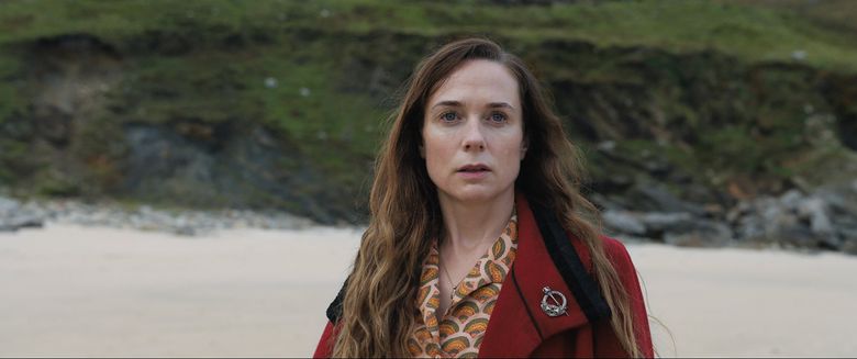 Kerry Condon stands on a beach, looking out of frame with an uneasy expression.