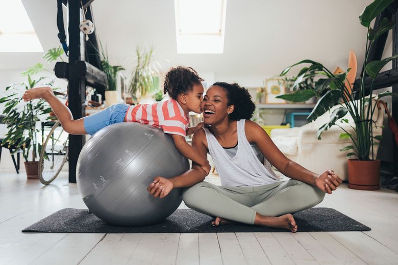 Woman sitting in a lotus pose on a mat, her little daughter on a pilates ball next to her kisses her cheek.