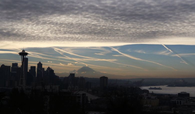 Contrails line the sky over Jet City and Mount Rainier. Contrails form when water vapor condenses around aerosols in jet exhaust. They create cirrus clouds that both trap heat (warming the earth) and reflect sunlight (cooling), with a net warming effect of uncertain magnitude. (Alan Berner / The Seattle Times, 2019)