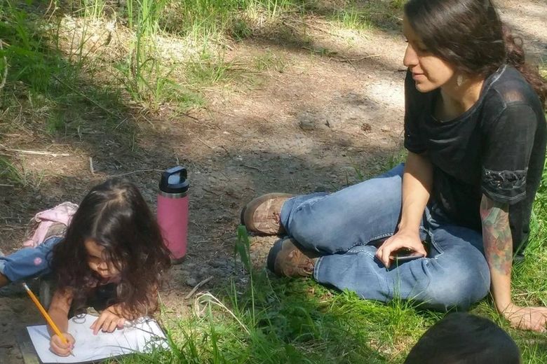 A woman in jeans and a T-shirt sits on the grass with a child who is coloring.