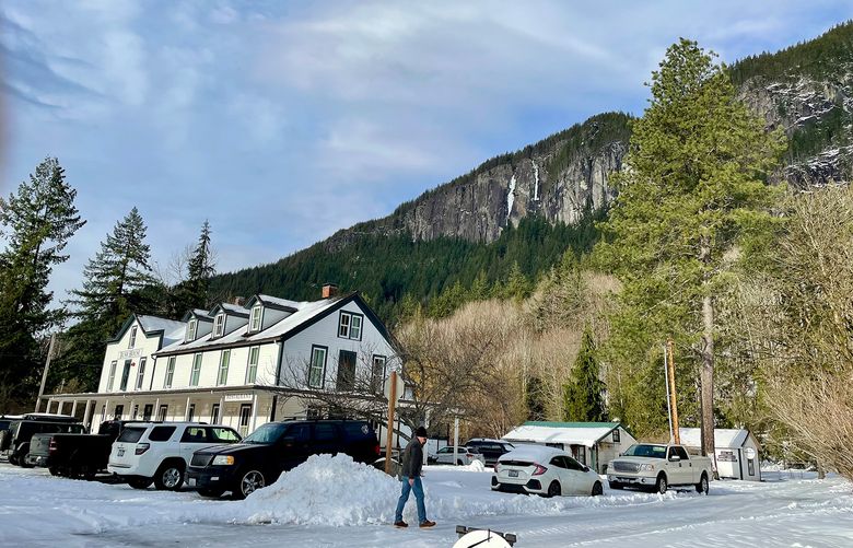 A man walks across a snow-covered path. A white inn lies in the background and evergreen trees stretch up the mountain in the distance.