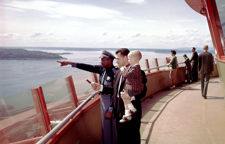 With West Seattle’s Admiral area and Alki Point as a backdrop, a Space Needle security guard points over low walls for a man and boy, both eating ice cream bars, on May 22, 1963. A small sign beyond the low glass panels warned, “Electrified System / Extreme Danger / Do Not Touch.”