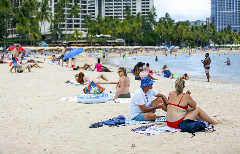 Brett Walsh and Emma Yates, bottom right, tourists from Australia, sit on Waikiki Beach in Honolulu, Monday, May 23, 2022. A COVID surge is under way that is starting to cause disruptions as schools wrap up for the year and Americans prepare for summer vacations. Case counts are as high as they’ve been since mid-February and those figures are likely a major undercount because of unreported home tests and asymptomatic infections. But the beaches beckoned and visitors have flocked to Hawaii, especially in recent months. (AP Photo/Caleb Jones) HICJ206