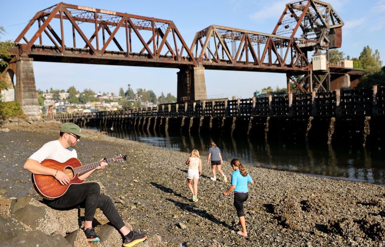 Ryan Dulmaine from Seattle writes a new song while sitting just west of the Ballard Locks under the The Salmon Bay Rail Bridge, also known as Bridge No. 4, at low tide while three girls beachcombing walk by on a sunny morning  in Seattle on Sunday, September 10, 2023.  The spot is “good inspiration” he says. “Its quiet. You can see the early morning activities. Its a nice place to clear your head and get thoughts out.”