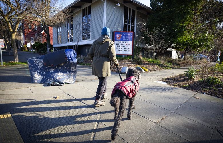 A dog walker passes a stack of possessions outside St. Luke’s Episcopal Church in Ballard, Thursday morning, March 7, 2024 in Seattle.