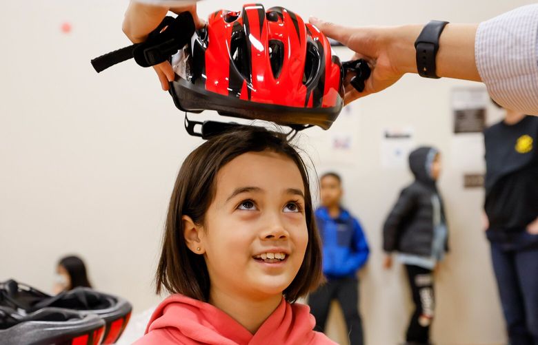Fourth grader Cecilia Mortensen looks up as Schuyler Peters places a bike helmet on her head at Meridian Park Elementary School Friday, March 22, 2024 in Shoreline, WA. Peters, a law student at Seattle U., organized the program to provide free bike helmets to kids. 226458