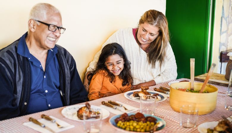 A woman laughs with a child at a dinner table alongside a senior man. There is an array of foods on the table.