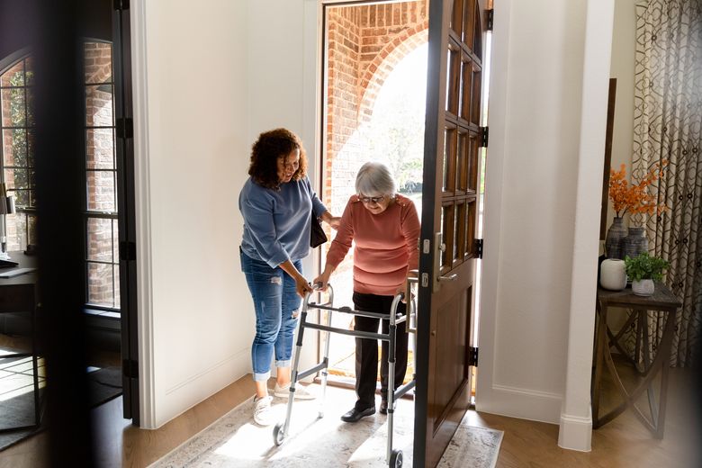 An adult woman helps a senior adult with a walker enter a home.