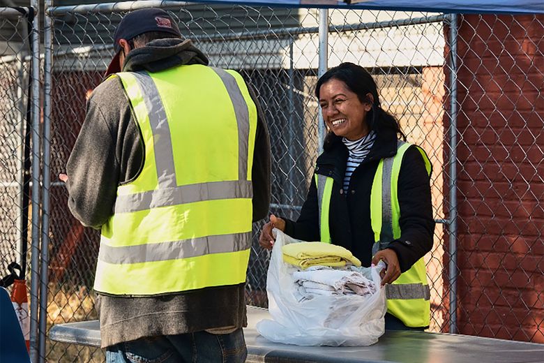 Two people wearing hi-vis vests stand outside next to a chain-link fence. One of them smiles, opening a plastic bag of folded towels.