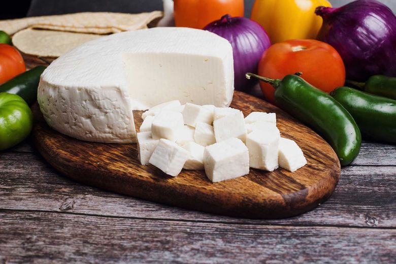 Cubes of Mexican white panela cheese sit in front of a round of the cheese, on a wooden cutting board, with jalapeno, tomato and red onion in the background.