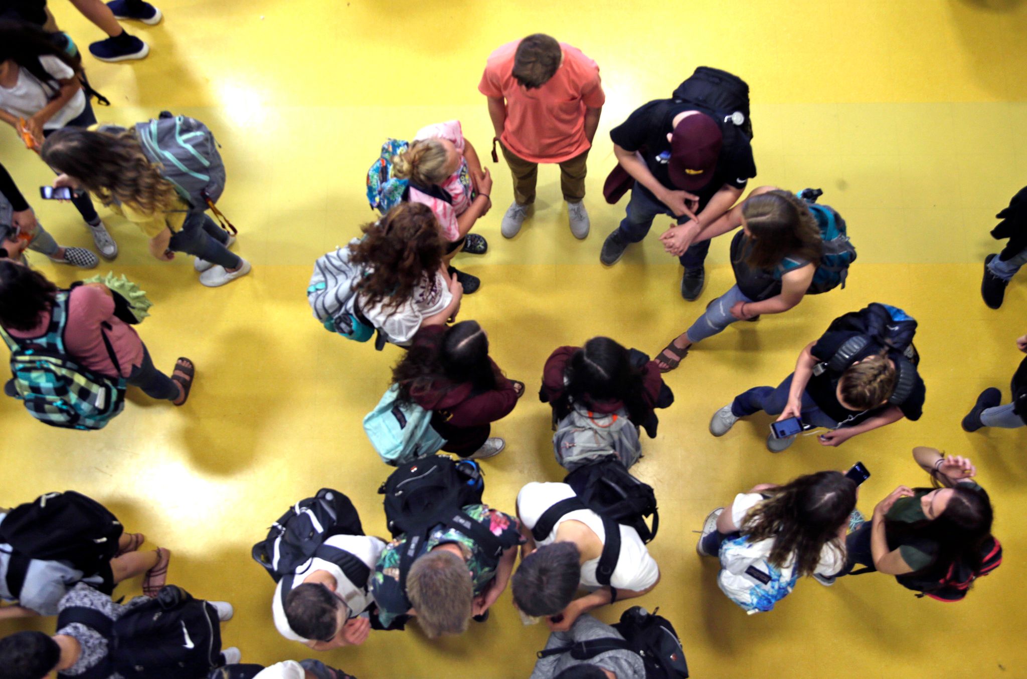 An overhead angle of several groups of students wearing backpacks, looking at phones, and talking to one another