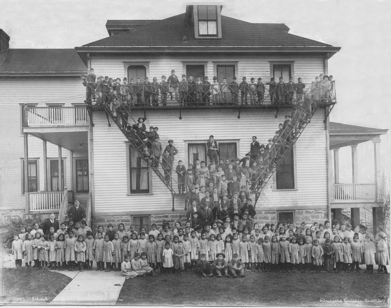 Фотография 1923 года, сделанная Дариусом Кинси недалеко от залива Тюлаллип, изображает студентов, стоящих у одного из общежитий Тюлаллипской интернатной школы. Школа закрылась в 1932 году и была одной из примерно 523 интернатов для коренных американцев, которые функционировали с 1801 по 1960-е годы в рамках федеральной и церковной политики ассимиляции. (Дариус Кинси / Культурный центр Хибулб)