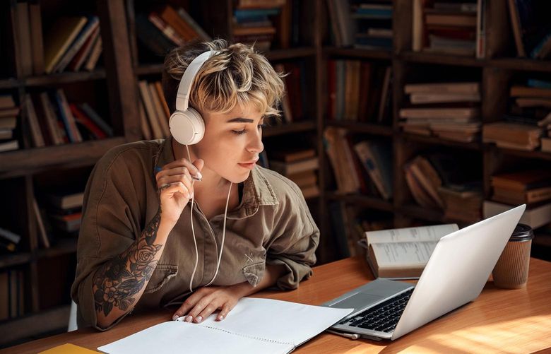 A concentrated and focused young adult woman wearing headphones works on her laptop at a desk in the library.