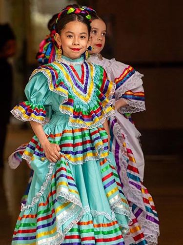 Two young Latina girls wearing colorful folklorico dresses prepare to dance.