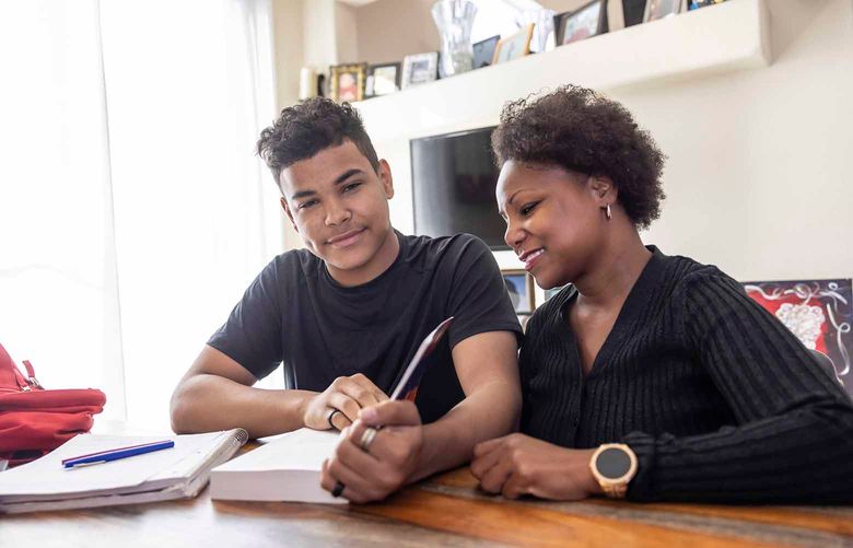 A high school student sits at a table with his mother, working on homework.