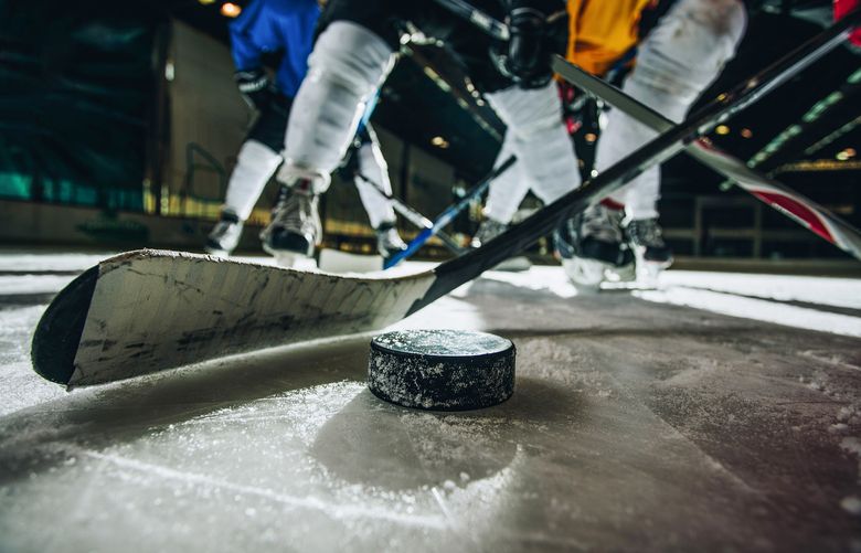 A close-up shot of a hockey puck on ice with two hockey players fighting for possession.