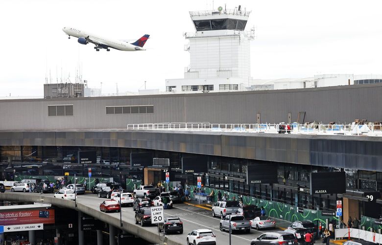 A plane from Delta Airlines flies past a ramp tower as cars drop off passengers at the departure gates at Seattle-Tacoma International Airport in SeaTac on Labor Day, Monday, September 2, 2024.