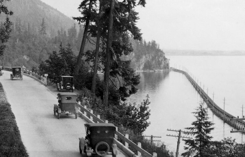 THEN: Bellingham photographer M.F. Jukes perched atop a 15-foot boulder over Chuckanut Drive circa 1920, looking south to Pigeon Point. The Everett-Bellingham Interurban trestle curves along Samish Bay. Unseen in this photo, Great Northern Railway tracks hug the shore. (Paul Dorpat Collection)