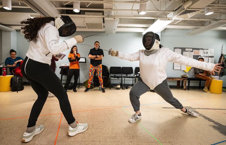 Staying within the lines of a lane-of-play called a “piste,” lead actors Anteia DeLaney, left, who plays the part of Mary Wallace, and Allison Renee, who plays the title role of Athena, run through a fencing scene during rehearsals for “Athena,” a theater collaboration between ArtsWest and fencing club Salle Auriol Seattle, Thursday, March 20, 2025, in West Seattle. In the background, from left, are director Kathryn Van Meter, stage manager Rebeca K. Hsia and fencing coach Sarah Lippai, from Salle Auriol Seattle.